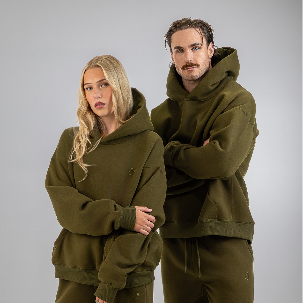 A man and a woman modeling matching olive green hoodies and sweatpants sets against a solid gray studio background.