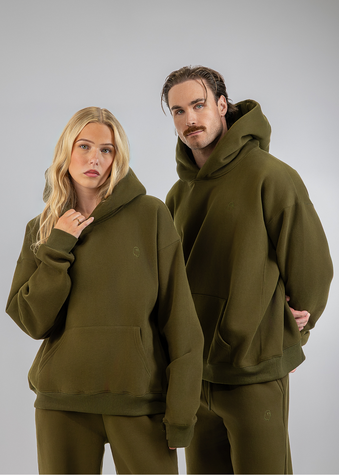 A man and a woman modeling matching olive green hoodies and sweatpants sets against a solid light gray studio background.