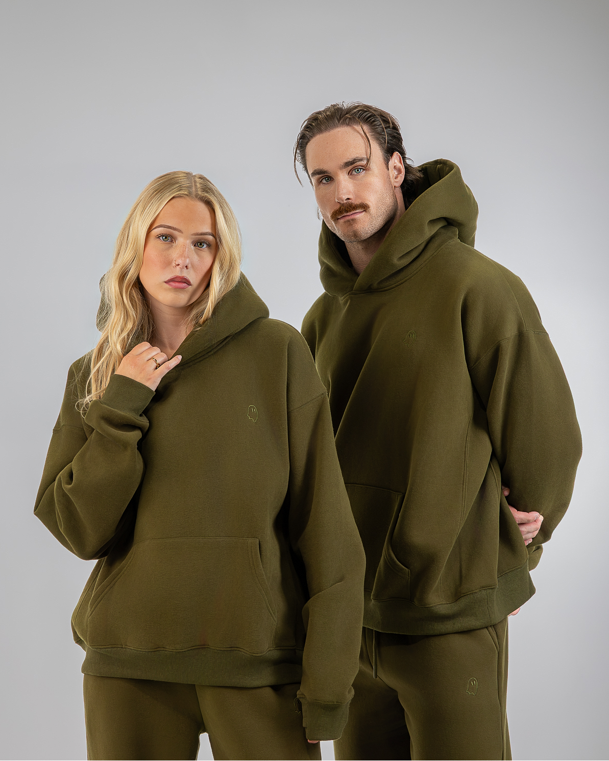 A man and a woman modeling matching olive green hoodies and sweatpants sets against a solid light gray studio background.