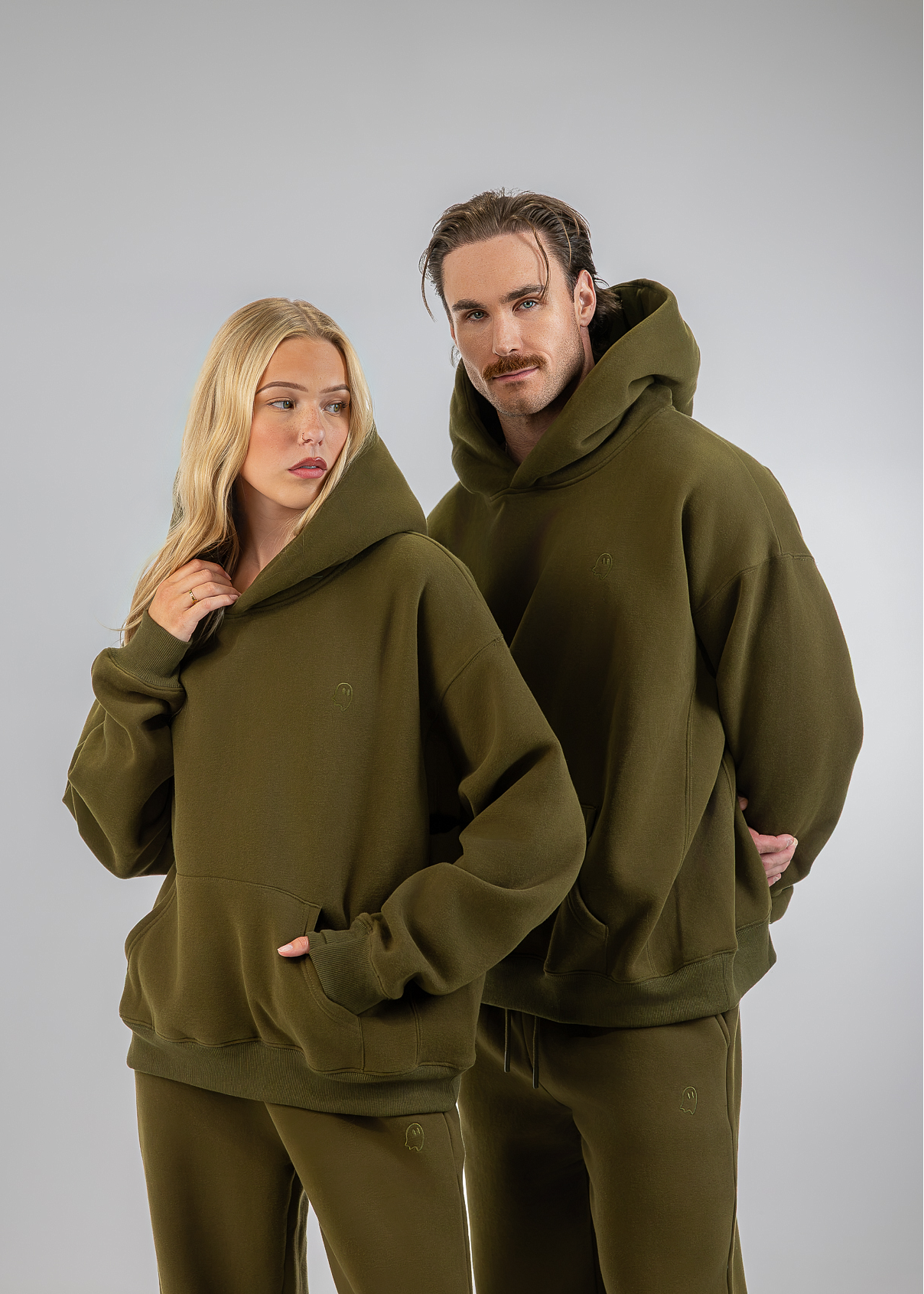 A man and a woman modeling matching olive green hooded sweatshirts and joggers against a plain light gray studio background.