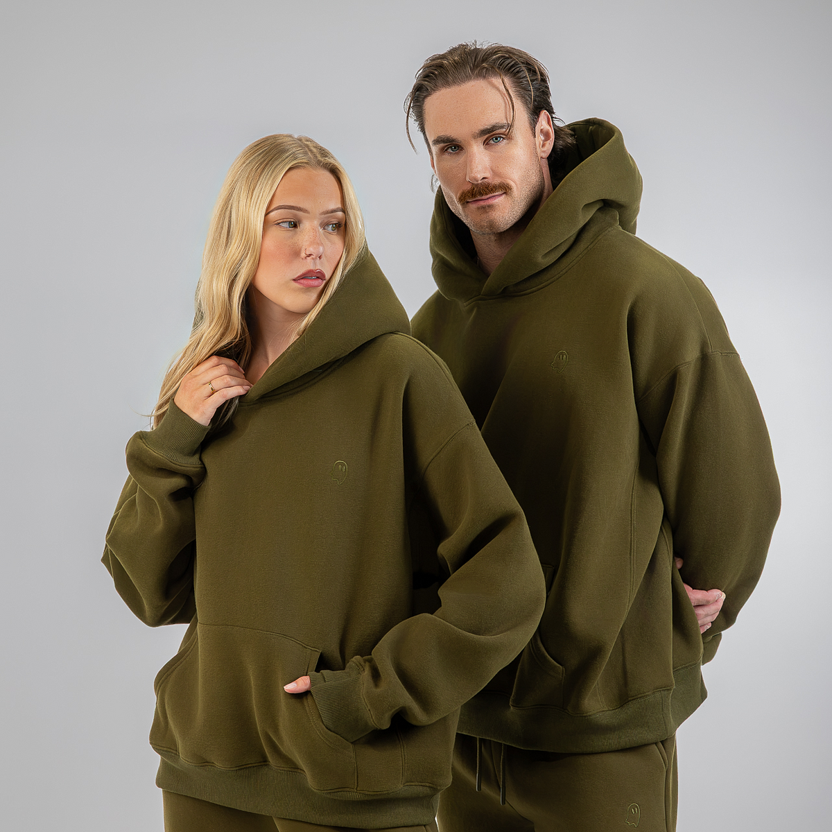 A man and a woman modeling matching olive green hooded sweatshirts and joggers against a plain light gray studio background.