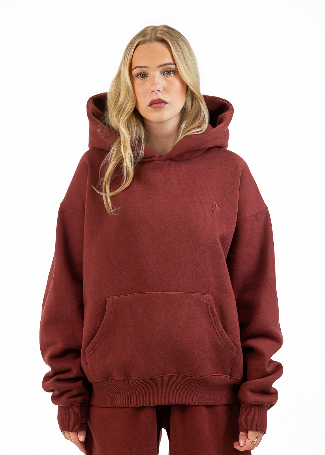 A blonde woman wearing a rust-brown pullover hoodie and matching sweatpants, standing against a solid white background.