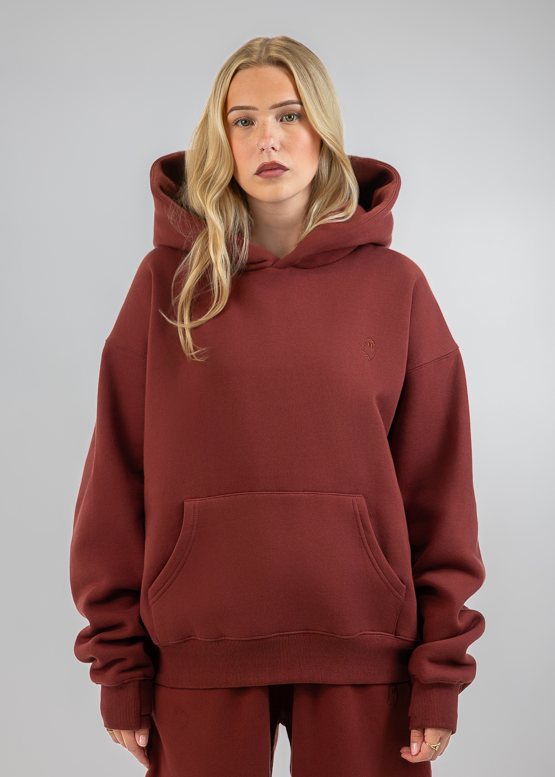 A blonde woman wearing an oversized rust-brown pullover hoodie with a kangaroo pocket and matching sweatpants, standing against a neutral gray studio background.