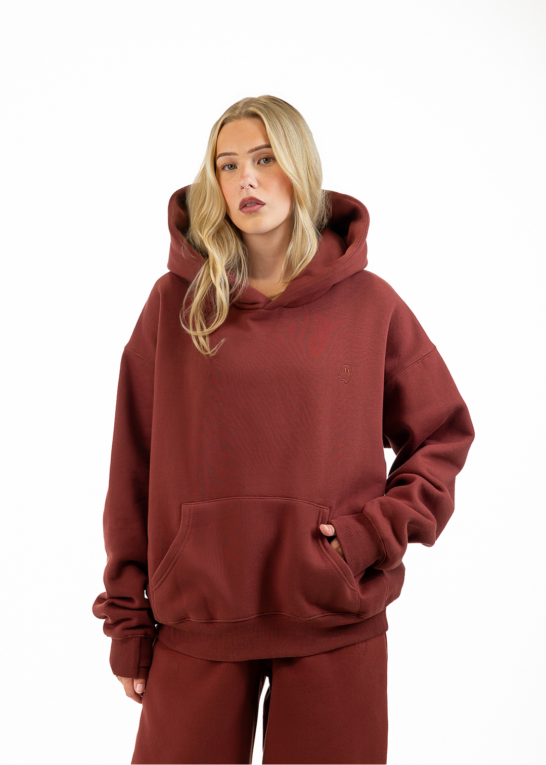 A blonde woman wearing an oversized rust-brown hoodie and matching sweatpants, standing against a plain white studio background.