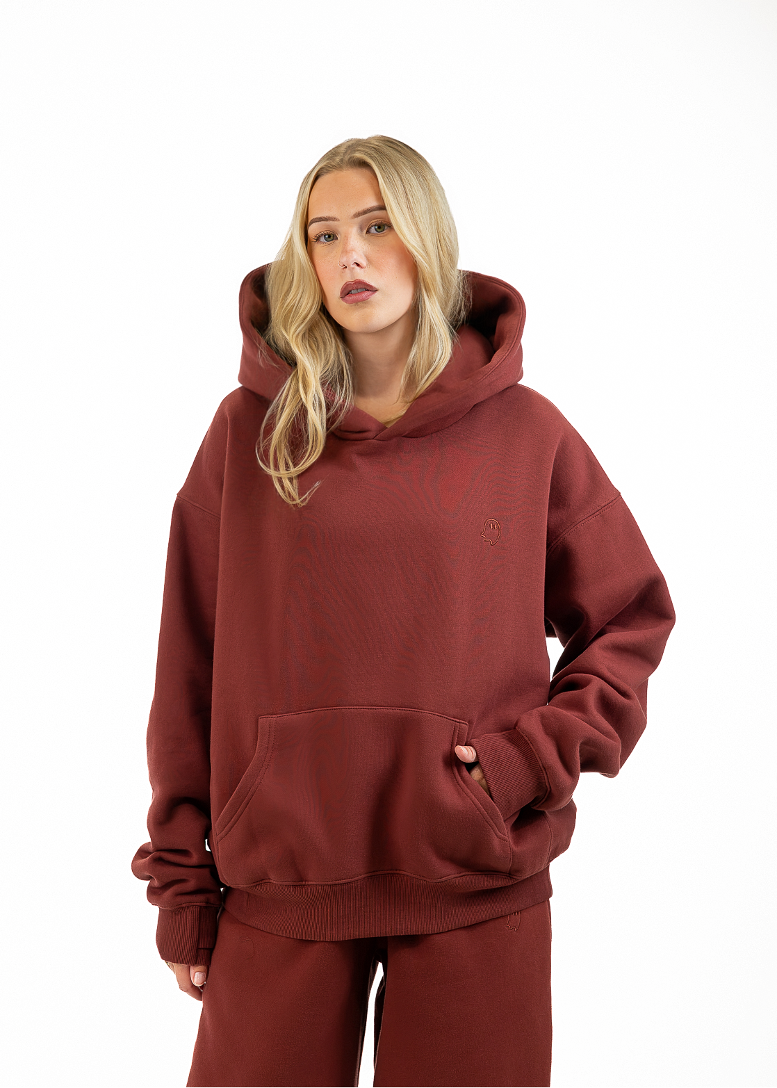 A blonde woman wearing an oversized rust-brown hoodie and matching sweatpants, standing against a plain white studio background.