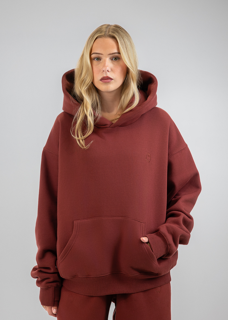 A blonde woman wearing an oversized rust-brown hooded sweatshirt and matching sweatpants, standing against a neutral gray studio background.