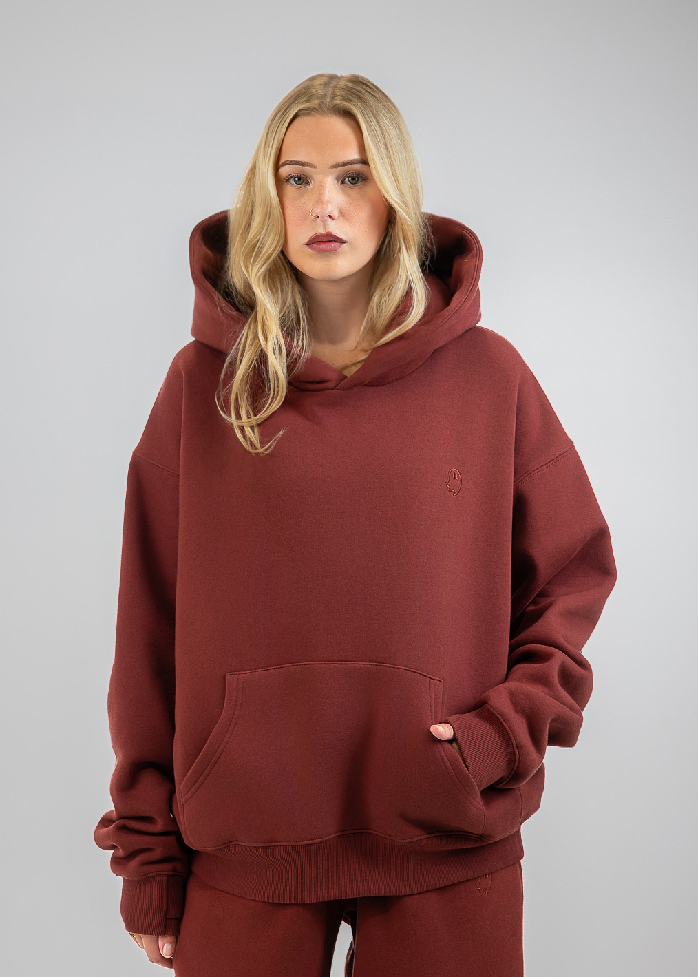 A blonde woman wearing an oversized rust-brown hooded sweatshirt and matching sweatpants, standing against a neutral gray studio background.