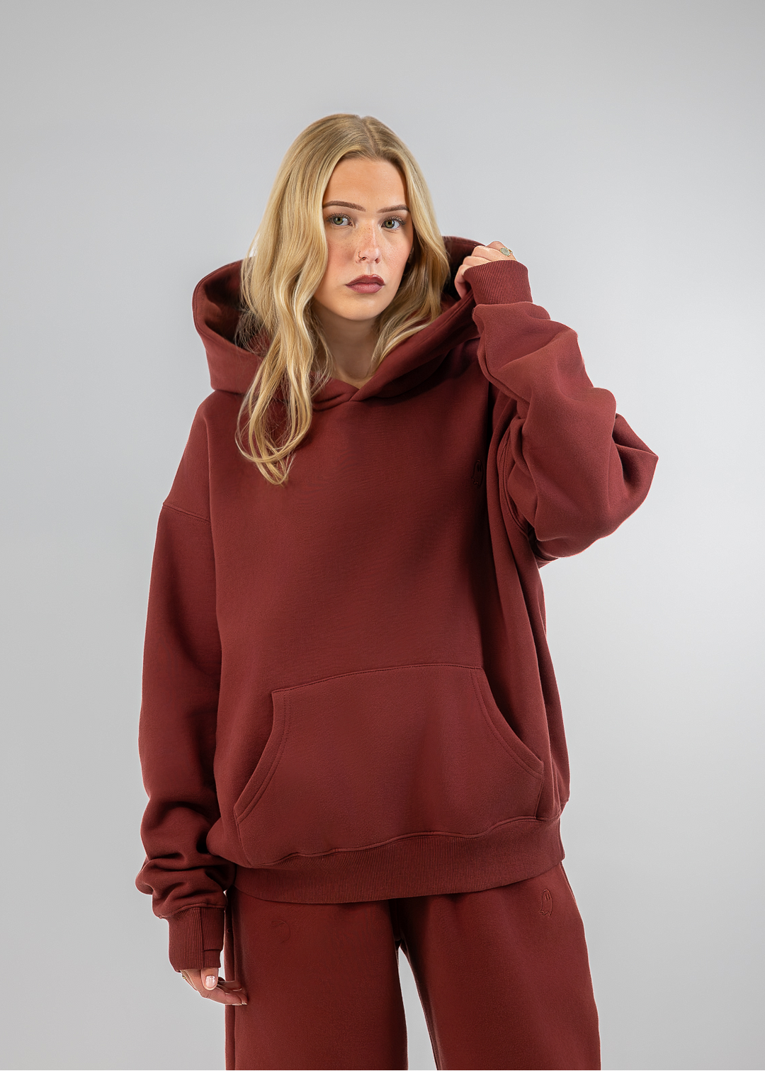 A woman modeling a matching rust-brown loungewear set consisting of an oversized pullover hoodie and wide-leg sweatpants, photographed against a neutral gray studio background.