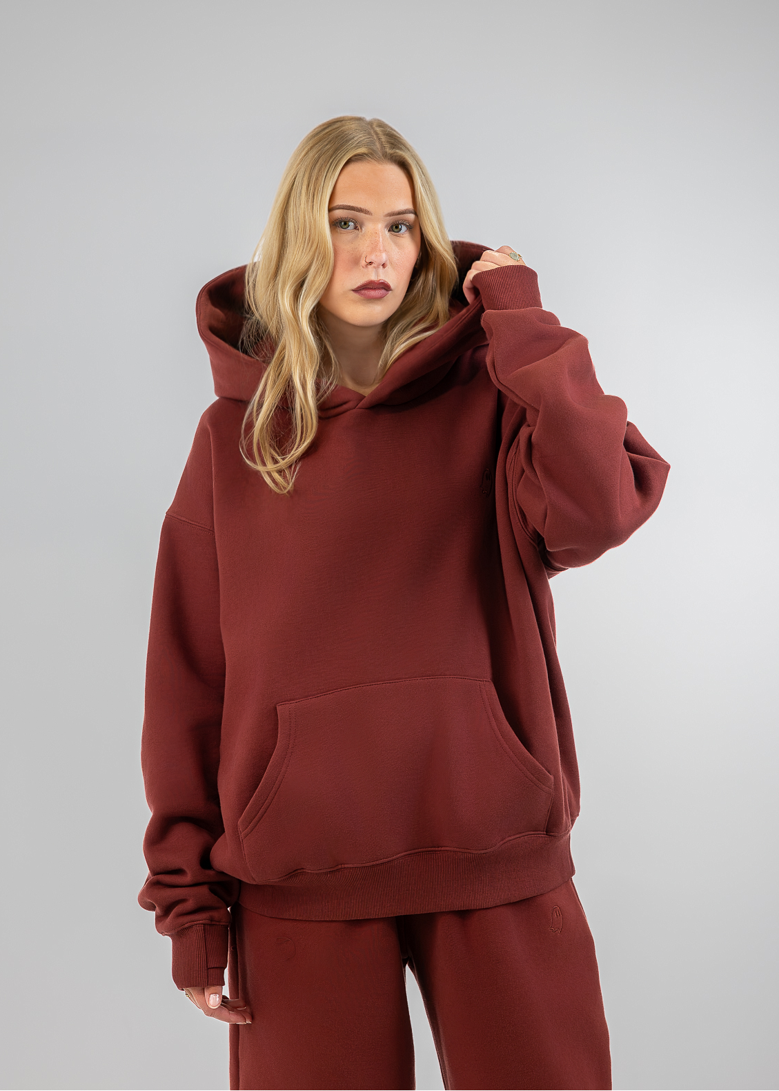A woman modeling a matching rust-brown loungewear set consisting of an oversized pullover hoodie and wide-leg sweatpants, photographed against a neutral gray studio background.