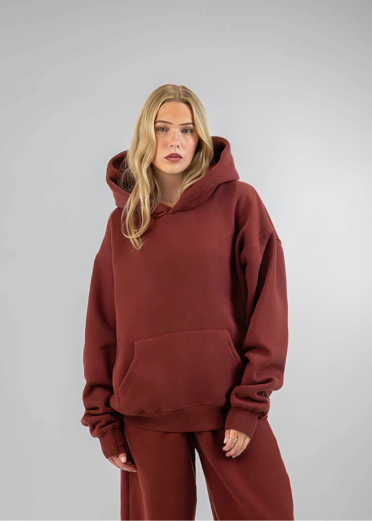 A woman modeling an oversized rust-brown hoodie and matching wide-leg sweatpants against a light gray studio background.