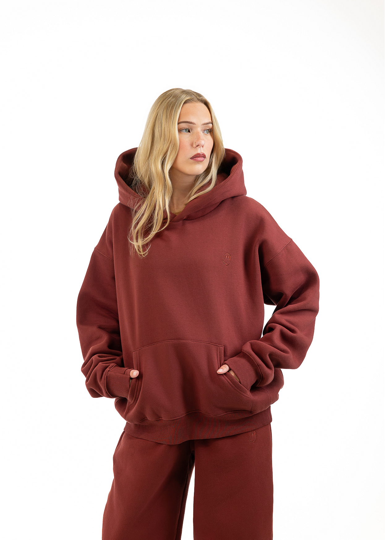 A woman modeling an oversized rust-brown hoodie and matching sweatpants against a plain white studio background.