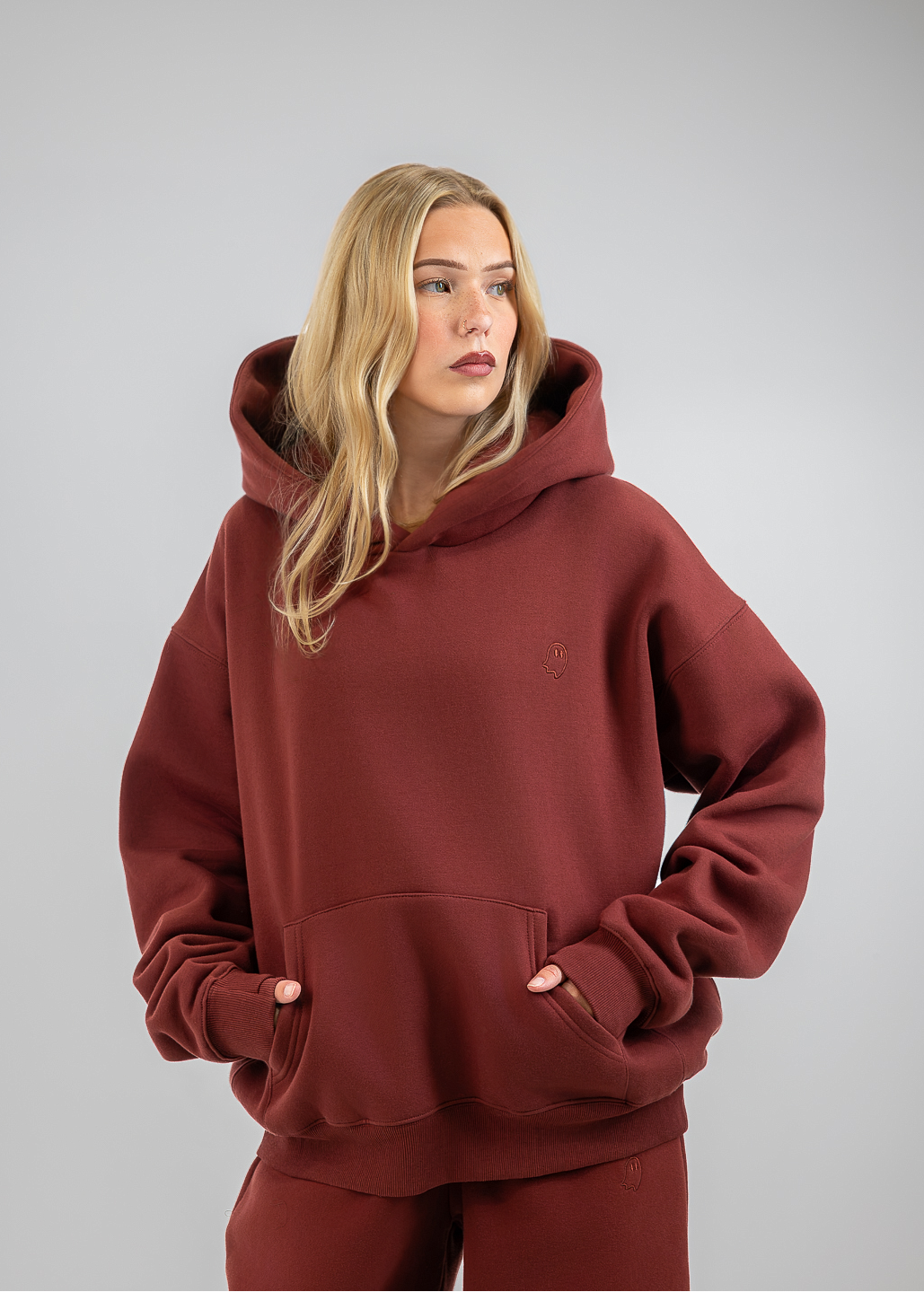A blonde woman wearing a matching rust-brown hooded sweatshirt and wide-leg sweatpants, standing against a neutral gray studio background.