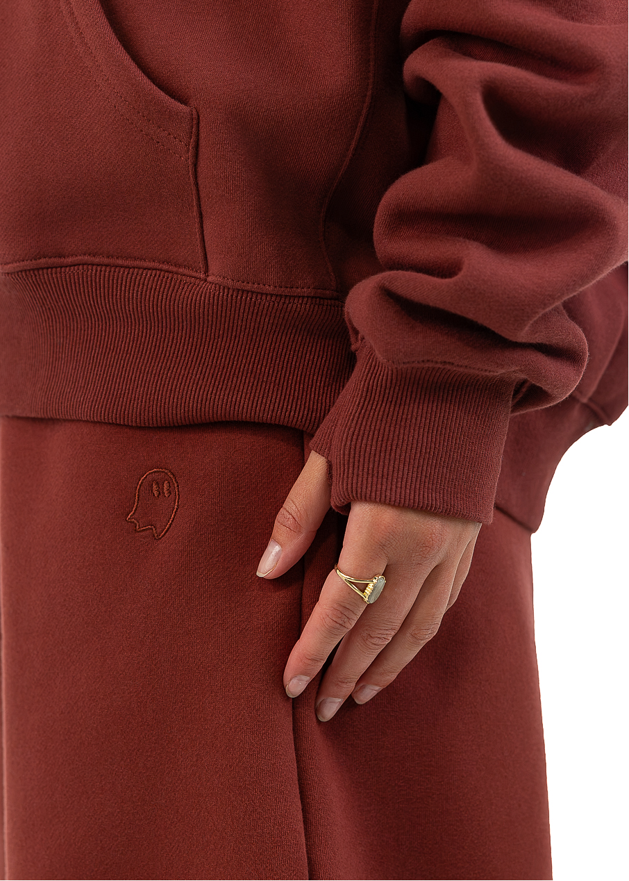 A close-up of a person wearing a matching rust-brown sweatshirt and sweatpants set, showing the side pocket, sleeve cuff, and hand resting on the pants.