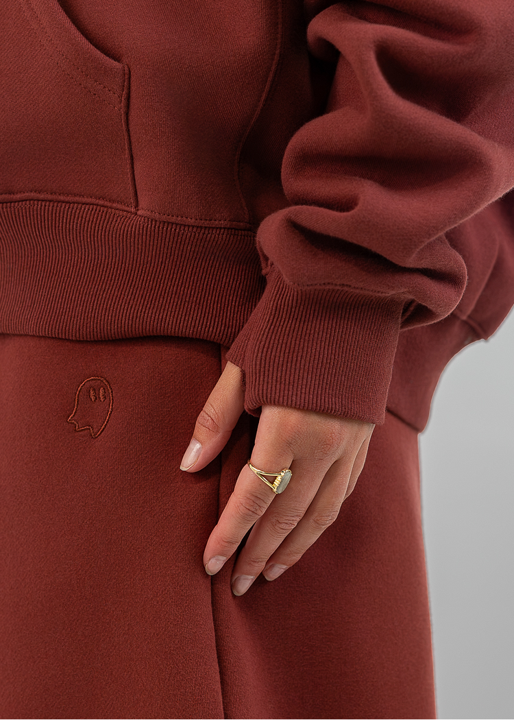 A close-up of a person wearing a rust-brown fleece hoodie and matching sweatpants, with one hand resting on the thigh. The person is wearing a gold ring.