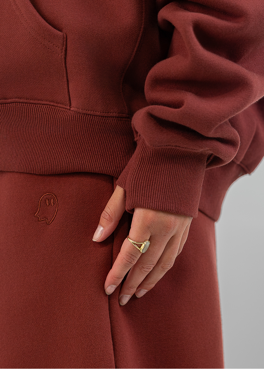 A close-up of a person wearing a rust-brown fleece hoodie and matching sweatpants, with one hand resting on the thigh. The person is wearing a gold ring.