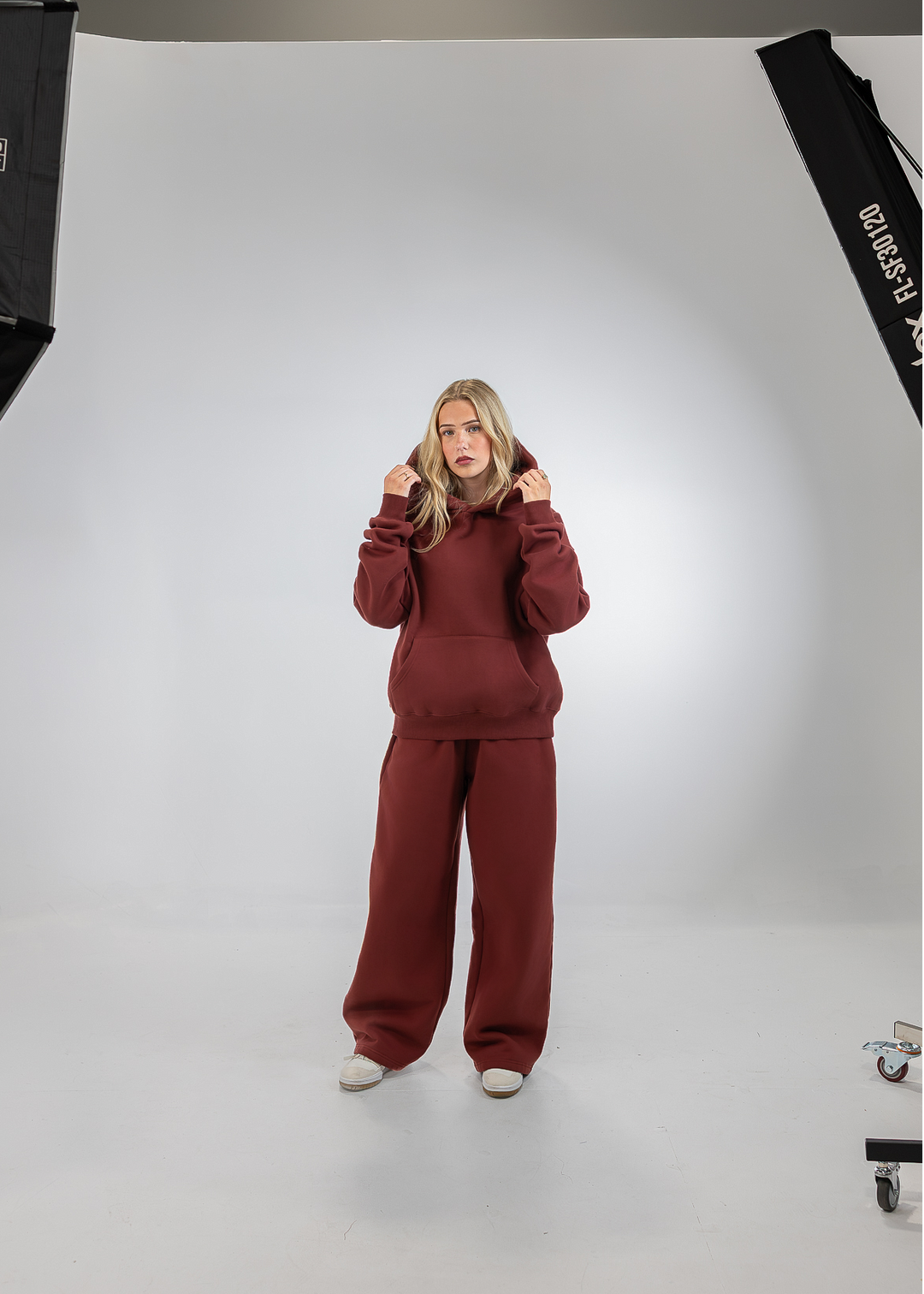 A woman stands in a studio wearing a matching burgundy sweatshirt and wide-leg sweatpants set. The photo is framed by softbox lights on both sides, with a white seamless backdrop.
