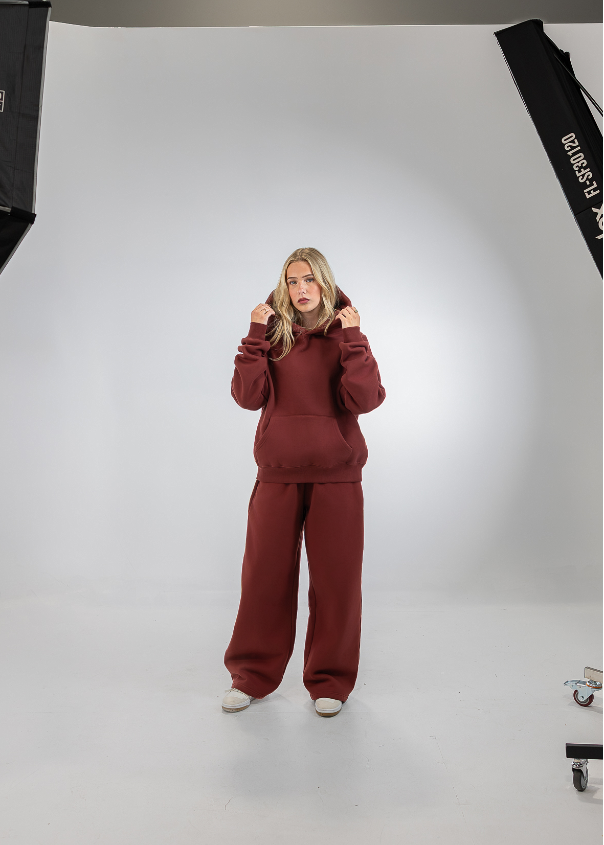 A woman stands in a studio wearing a matching burgundy sweatshirt and wide-leg sweatpants set. The photo is framed by softbox lights on both sides, with a white seamless backdrop.