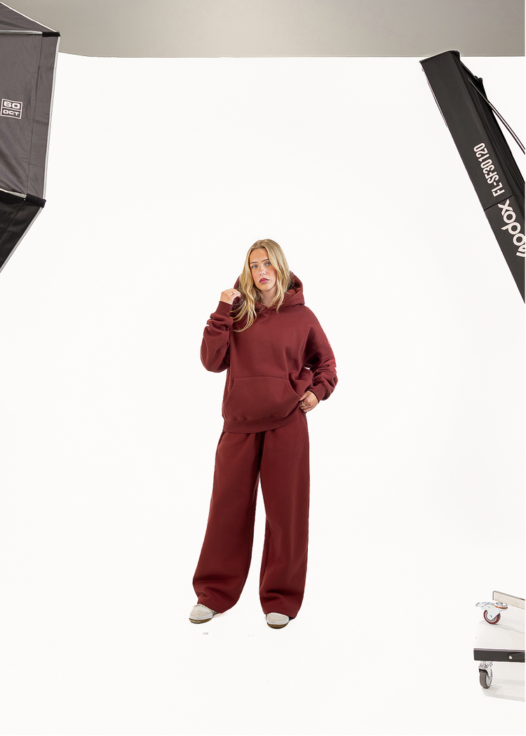 A woman modeling a matching burgundy lounge set consisting of a pullover hoodie and wide-leg sweatpants, standing against a white studio backdrop with softbox lights visible on the left and right.
