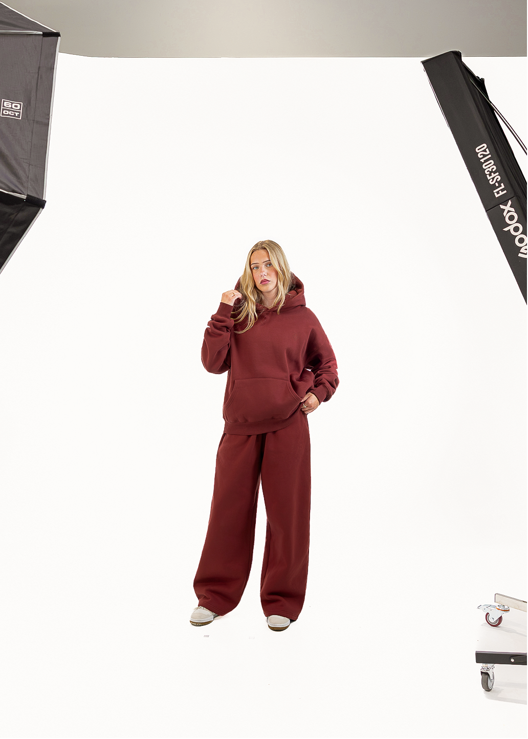 A woman modeling a matching burgundy lounge set consisting of a pullover hoodie and wide-leg sweatpants, standing against a white studio backdrop with softbox lights visible on the left and right.