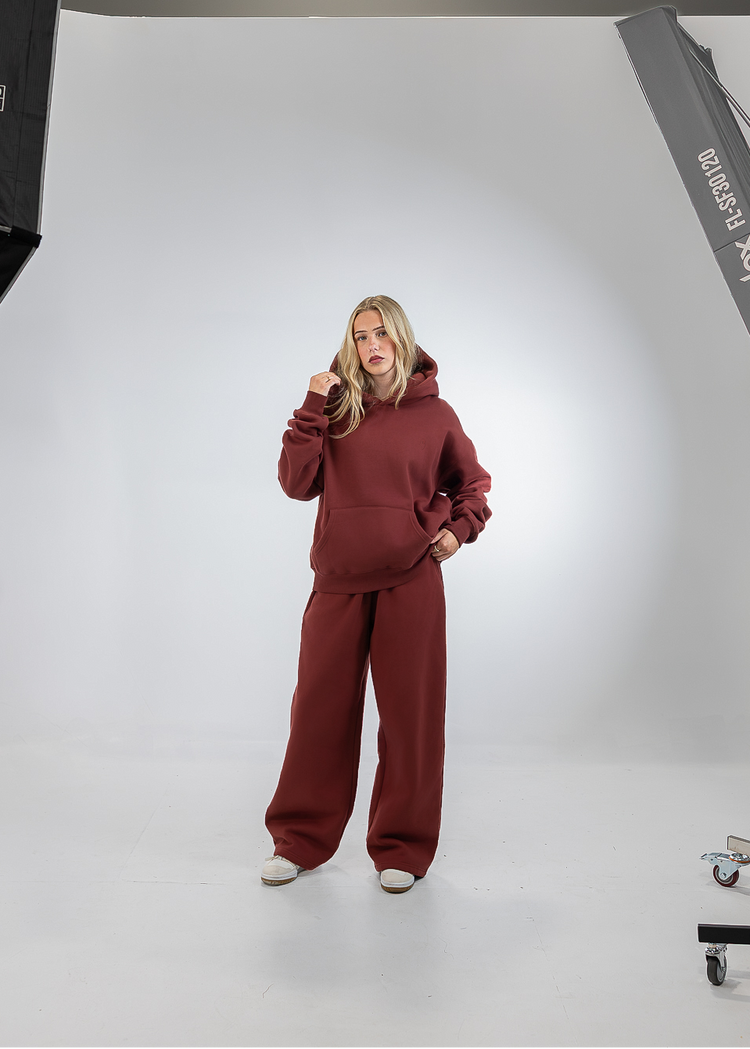 A woman modeling a matching burgundy lounge set consisting of a pullover hoodie and wide-leg sweatpants, standing in a white studio setting with softbox lights visible on the left and right.