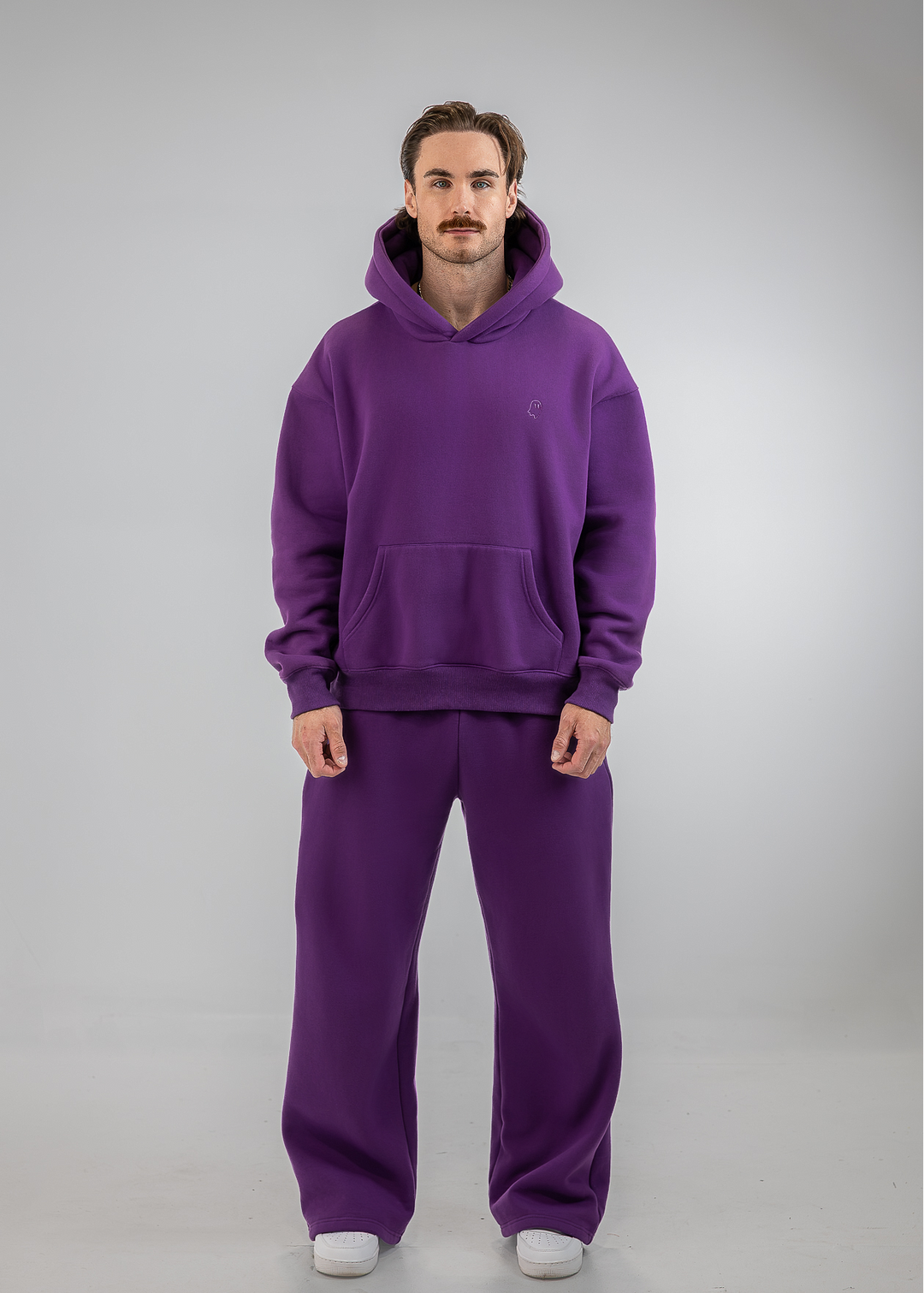 A man modeling a matching purple sweatshirt and sweatpants set against a light gray studio background.