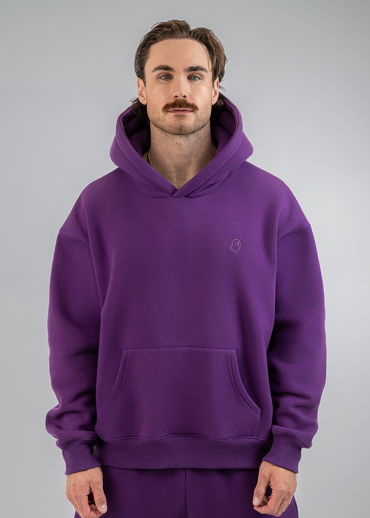 A man wearing a matching purple hoodie and sweatpants set, standing against a neutral gray studio background.