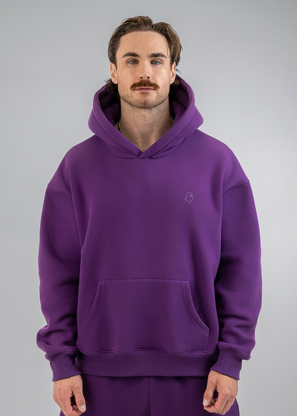 A man wearing a matching purple hoodie and sweatpants set, standing against a neutral gray studio background.