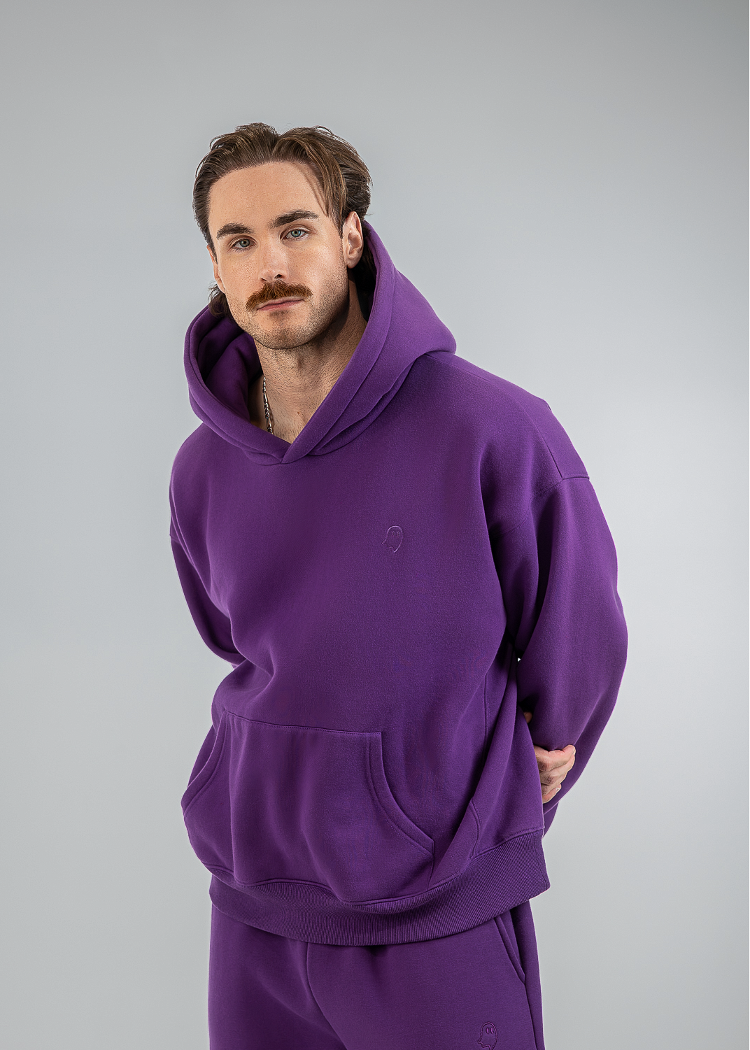 A man modeling a matching purple hoodie and sweatpants set against a plain light gray studio background.