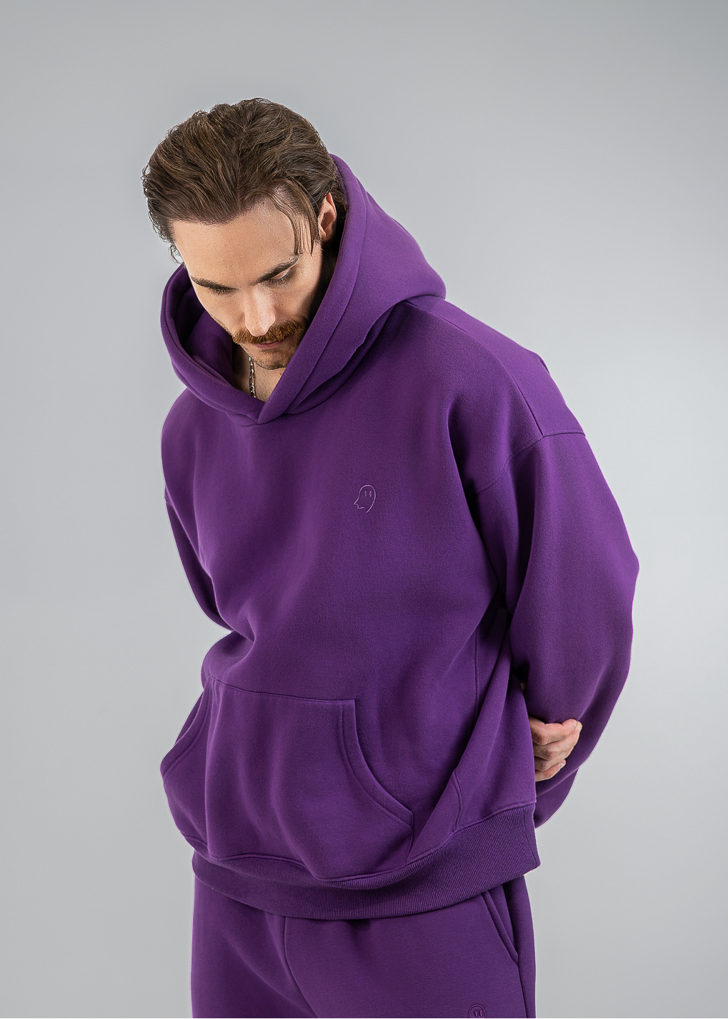 A man wearing a matching purple hoodie and sweatpants set, photographed against a neutral gray studio background.