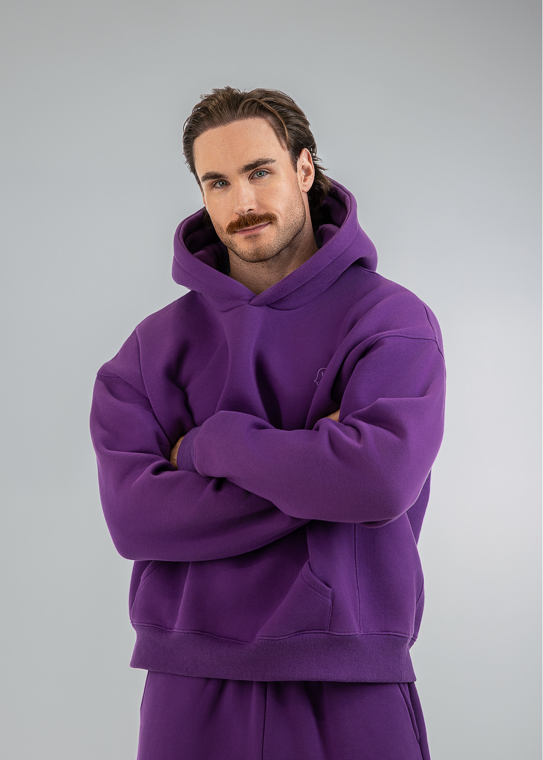 A man wearing a matching purple hoodie and sweatpants set, standing with arms crossed against a light gray studio background.