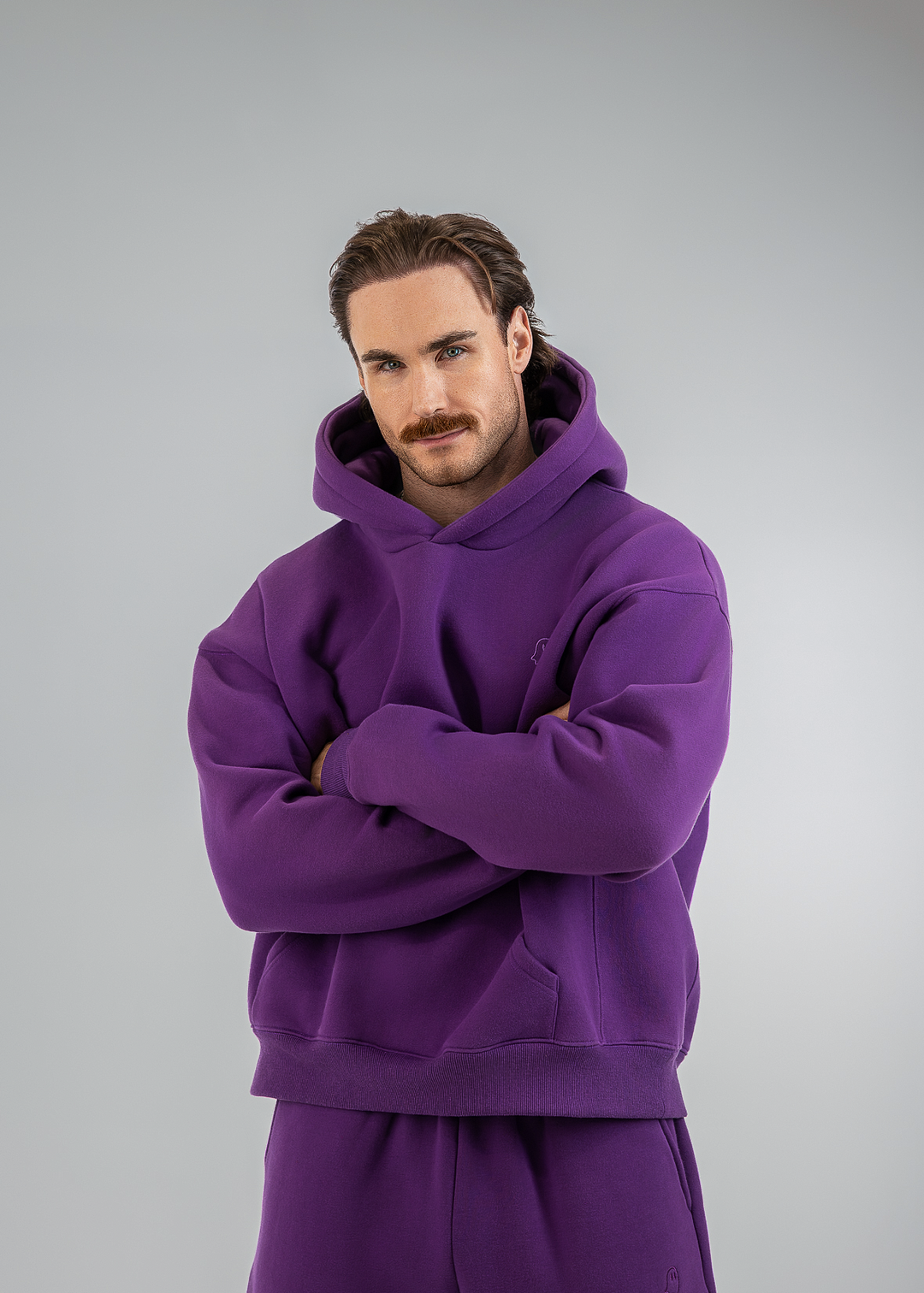 A man wearing a matching purple hoodie and sweatpants set, standing with arms crossed against a light gray studio background.
