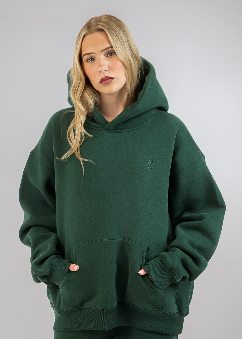 A blonde woman wearing an oversized dark green hoodie and matching green sweatpants, standing against a neutral gray studio background.