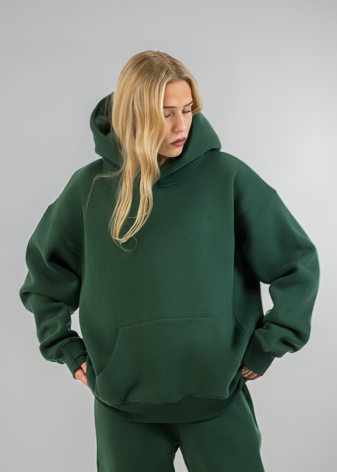 A blonde woman wearing an oversized dark green hoodie and matching sweatpants, standing against a neutral light gray studio background.