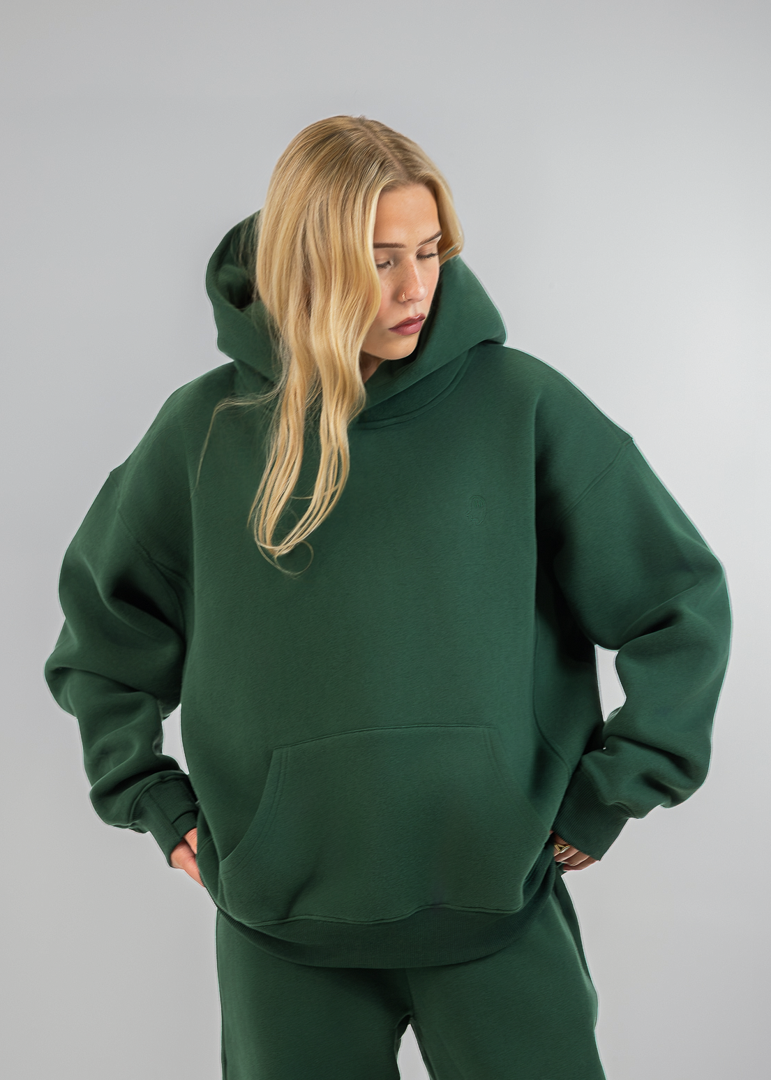 A blonde woman wearing an oversized dark green hoodie and matching sweatpants, standing against a neutral light gray studio background.