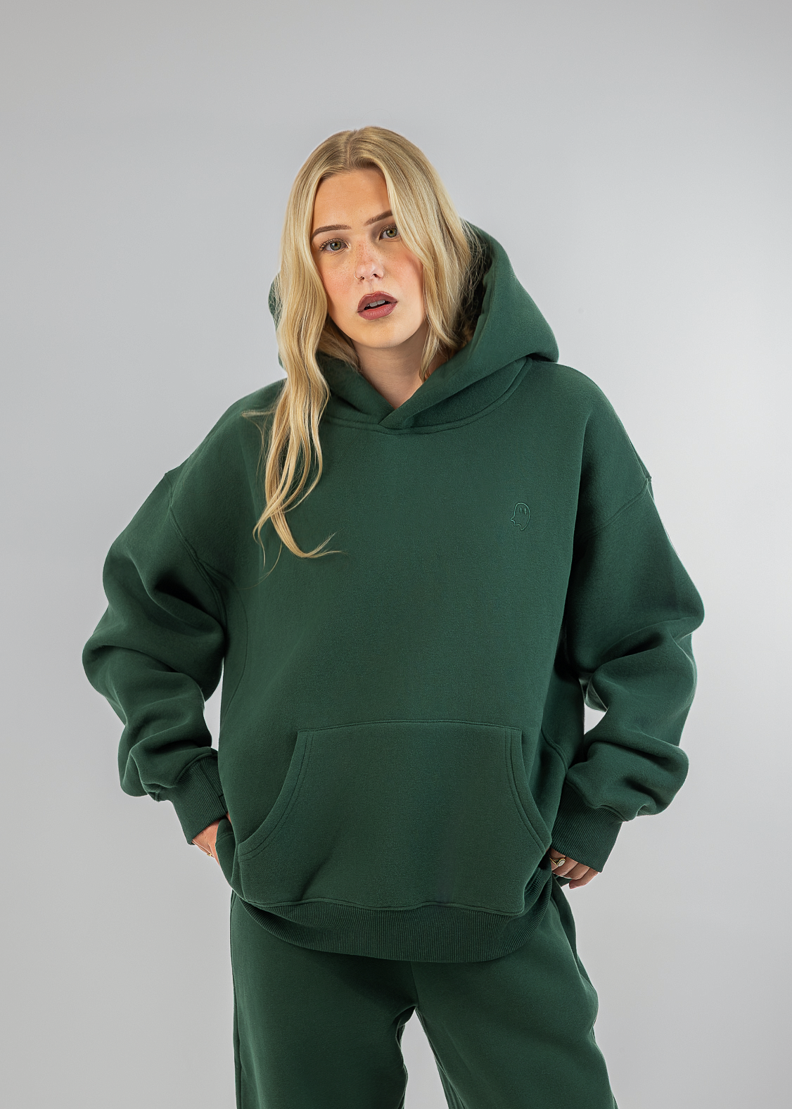 A woman modeling an oversized dark green hoodie and matching sweatpants against a plain light gray studio background.