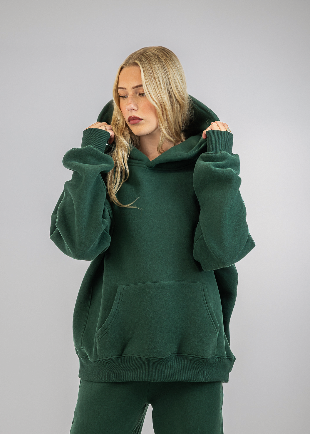 A woman wearing an oversized dark green hoodie and matching sweatpants, posing against a neutral gray studio background.