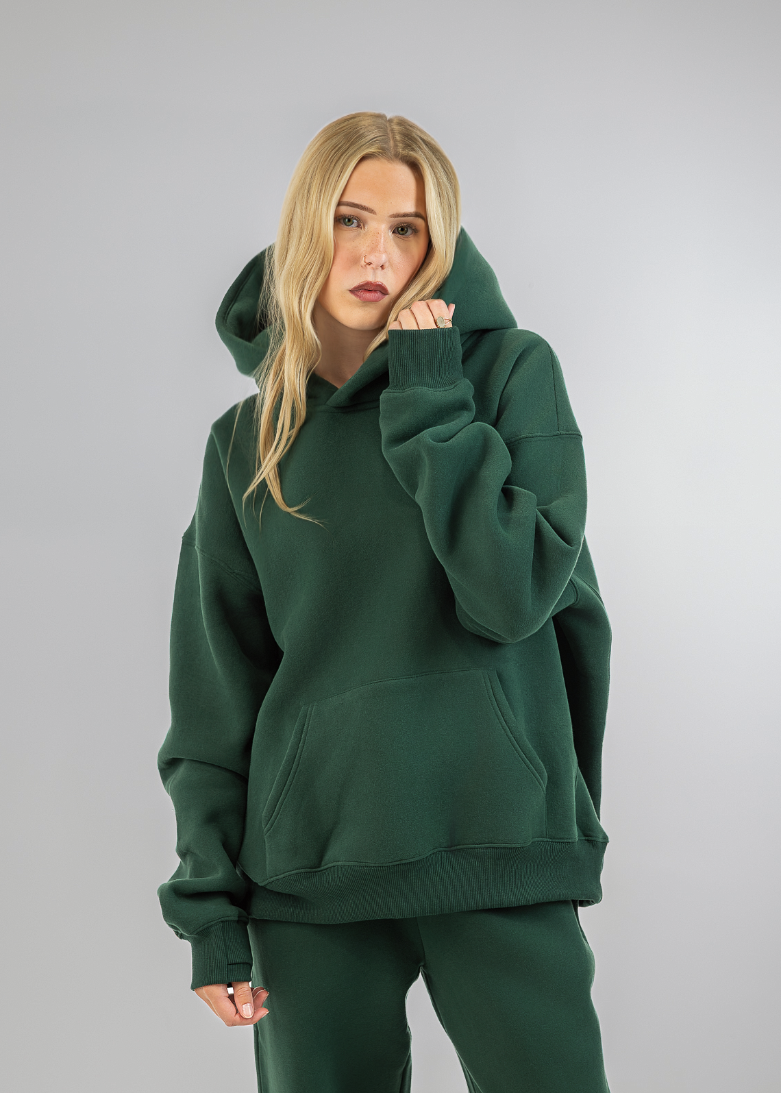 A blonde model wearing an oversized dark green hoodie and matching sweatpants, posing against a neutral light gray studio background.