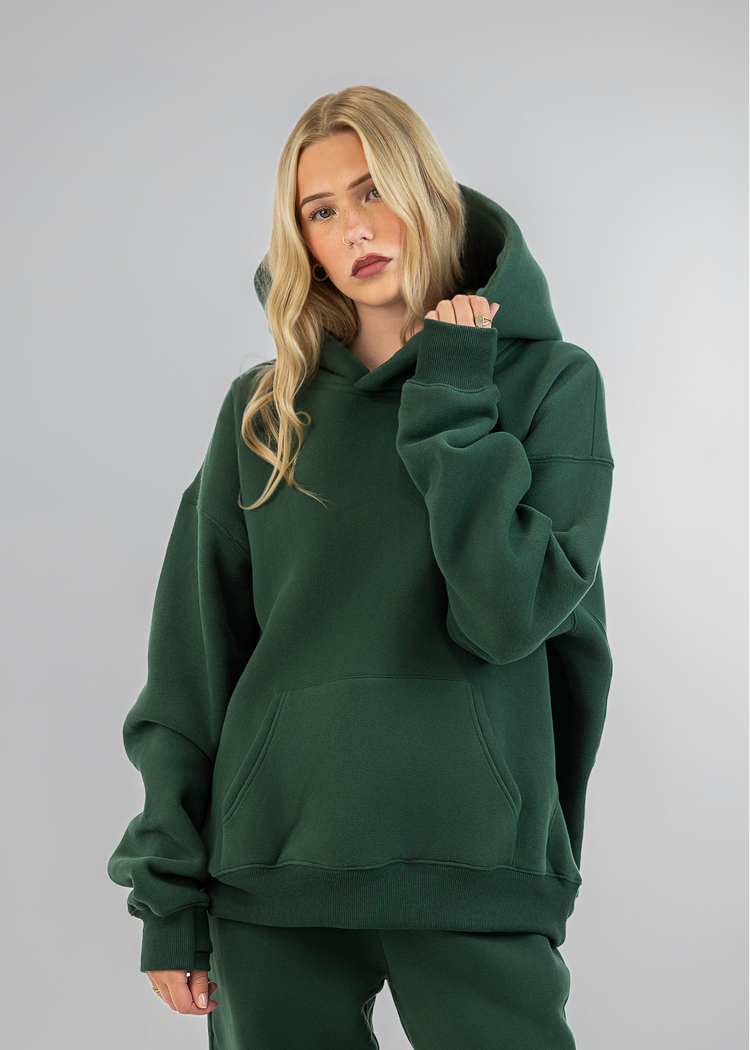 A blonde model wearing an oversized dark green hoodie and matching sweatpants, standing against a neutral gray studio background.