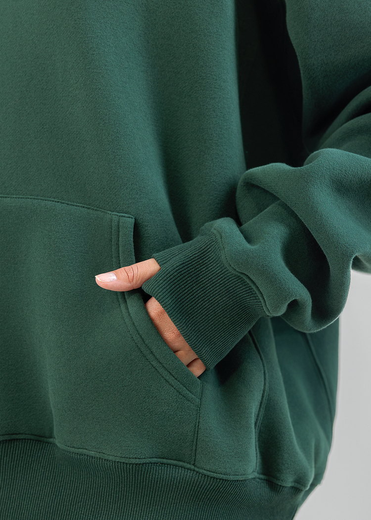 Close-up of a person wearing a dark green sweatshirt and matching sweatpants, with one hand tucked into the front pocket. The photo is cropped to show the torso and sleeves against a plain light gray background.