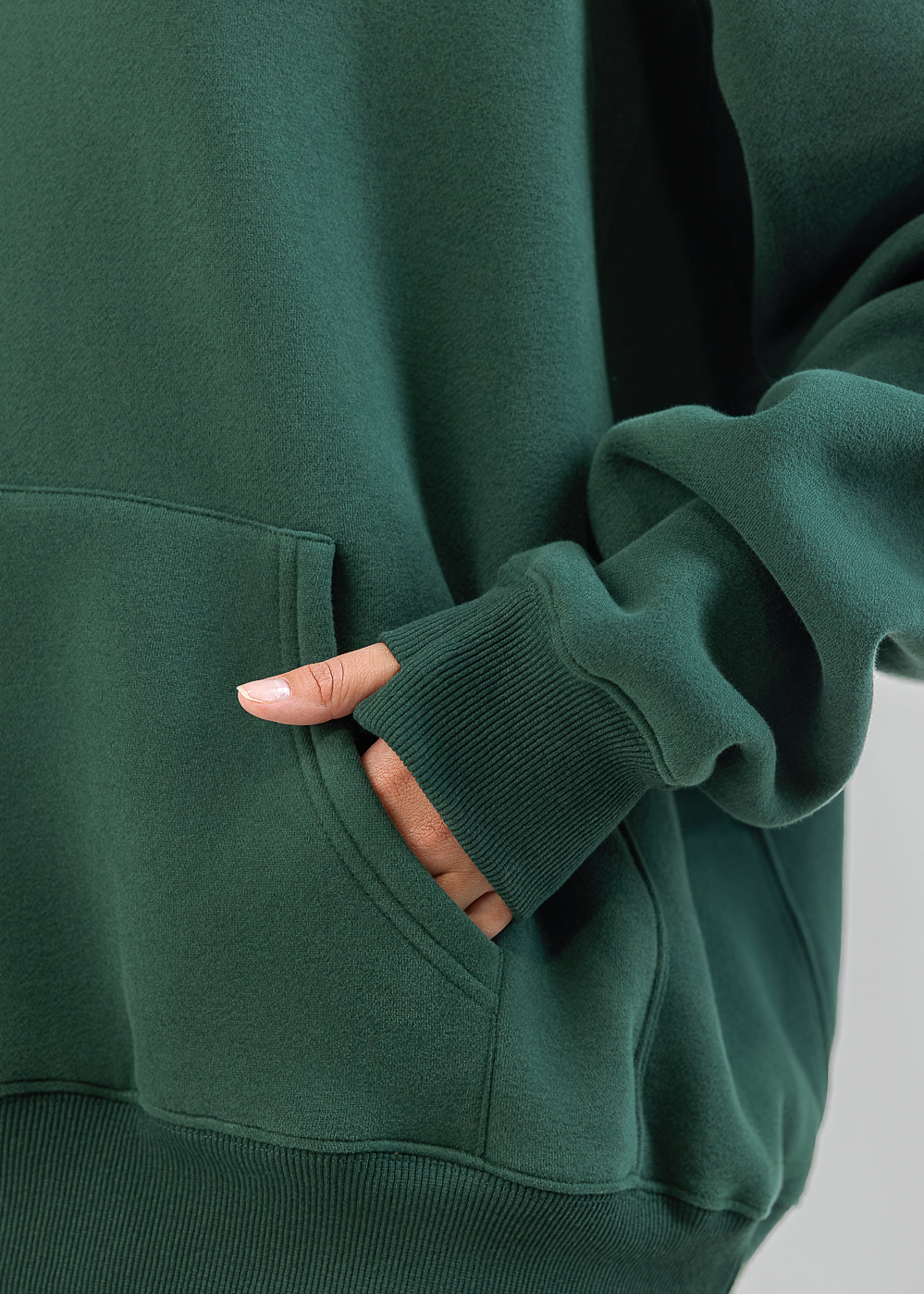Close-up of a person wearing a dark green sweatshirt and matching sweatpants, with one hand tucked into the front pocket. The photo is cropped to show the torso and sleeves against a plain light gray background.