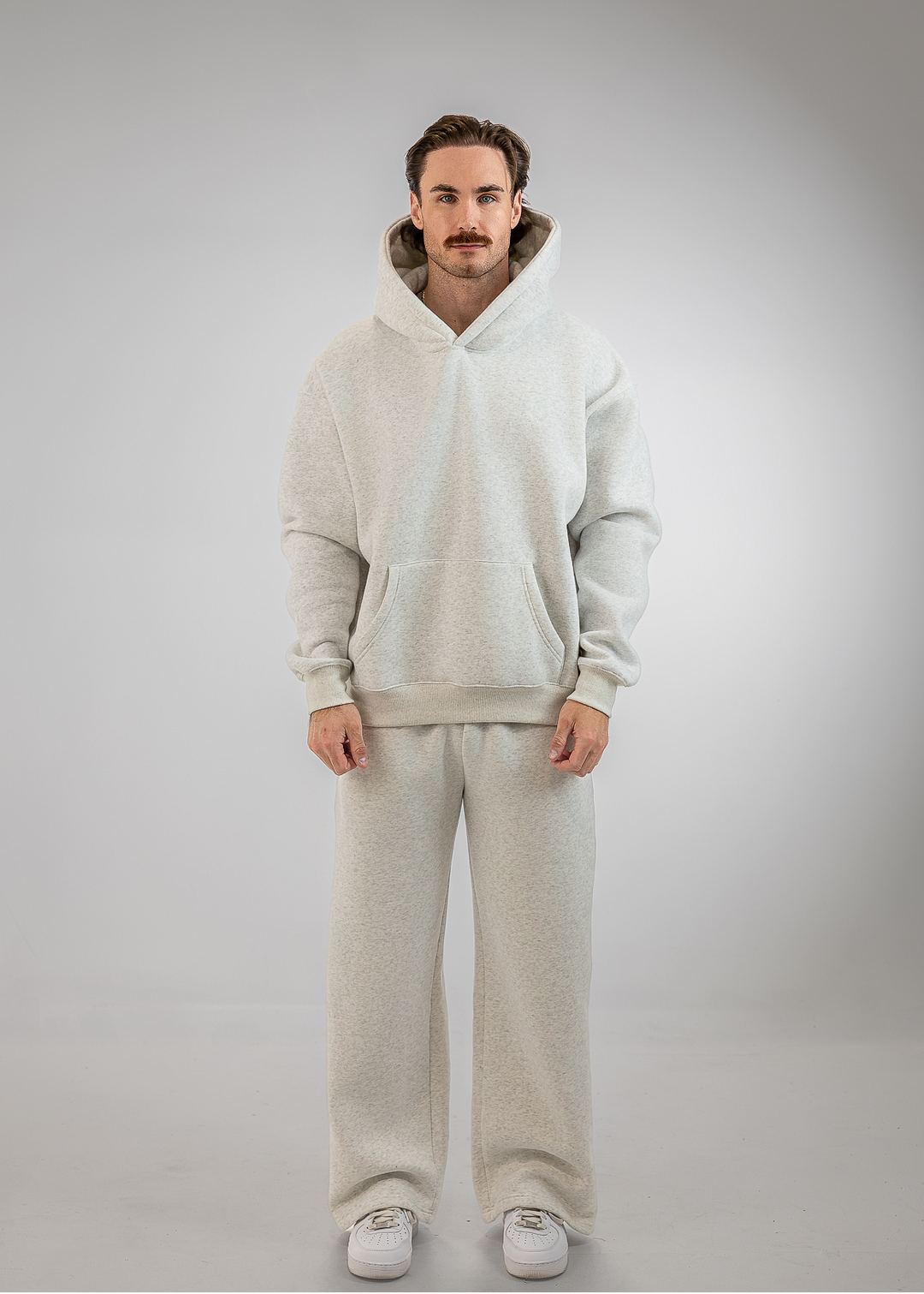 A man wearing a matching light beige fleece hoodie and sweatpants set with white sneakers, standing against a neutral gray studio background.