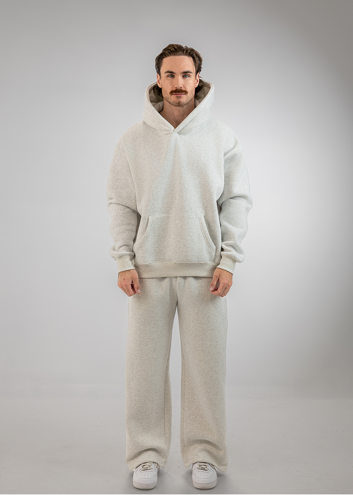 A man wearing a matching light beige fleece hoodie and sweatpants set with white sneakers, standing against a neutral gray studio background.