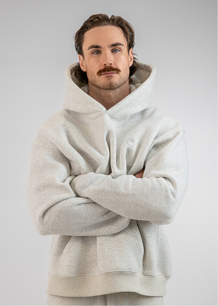 A man wearing a light gray oversized hoodie and matching sweatpants, standing with his arms crossed against a plain light gray studio background.