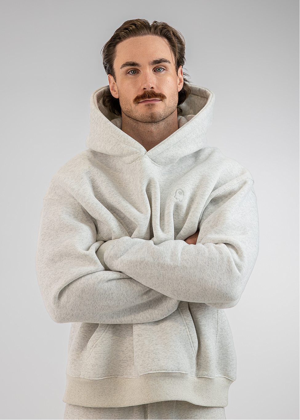 A man wearing a light gray oversized hoodie and matching sweatpants, standing with his arms crossed against a plain light gray studio background.