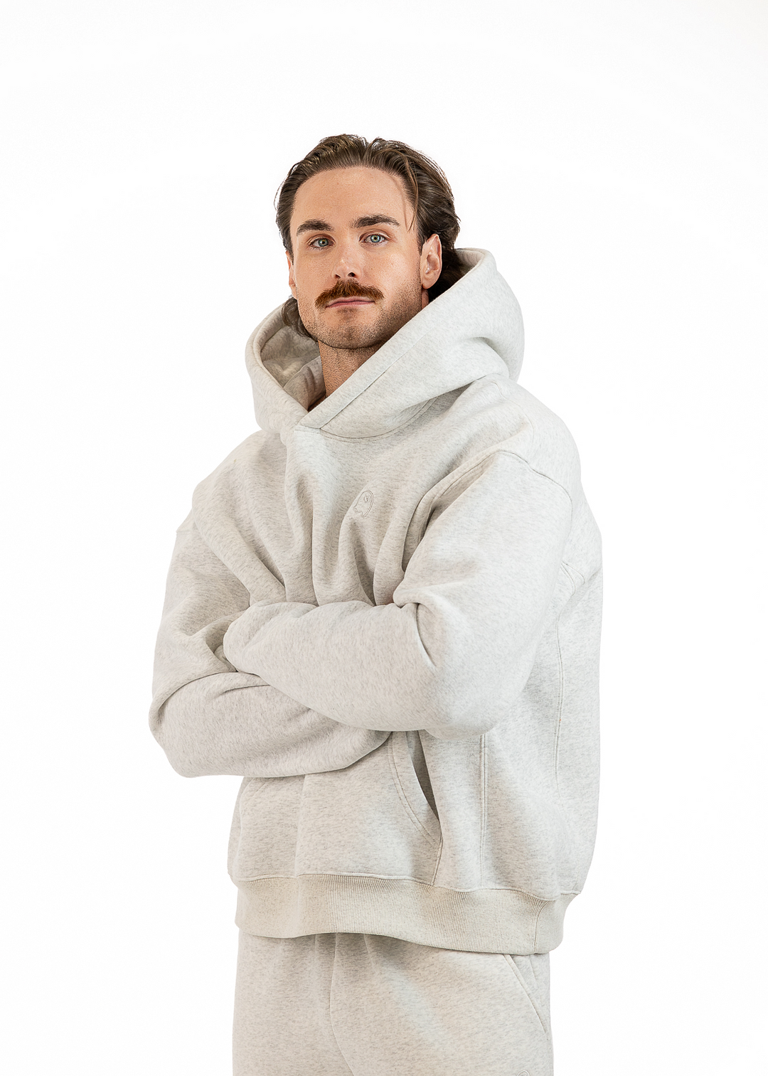 A man wearing a light gray oversized hoodie and matching sweatpants, standing with arms crossed against a plain white background.