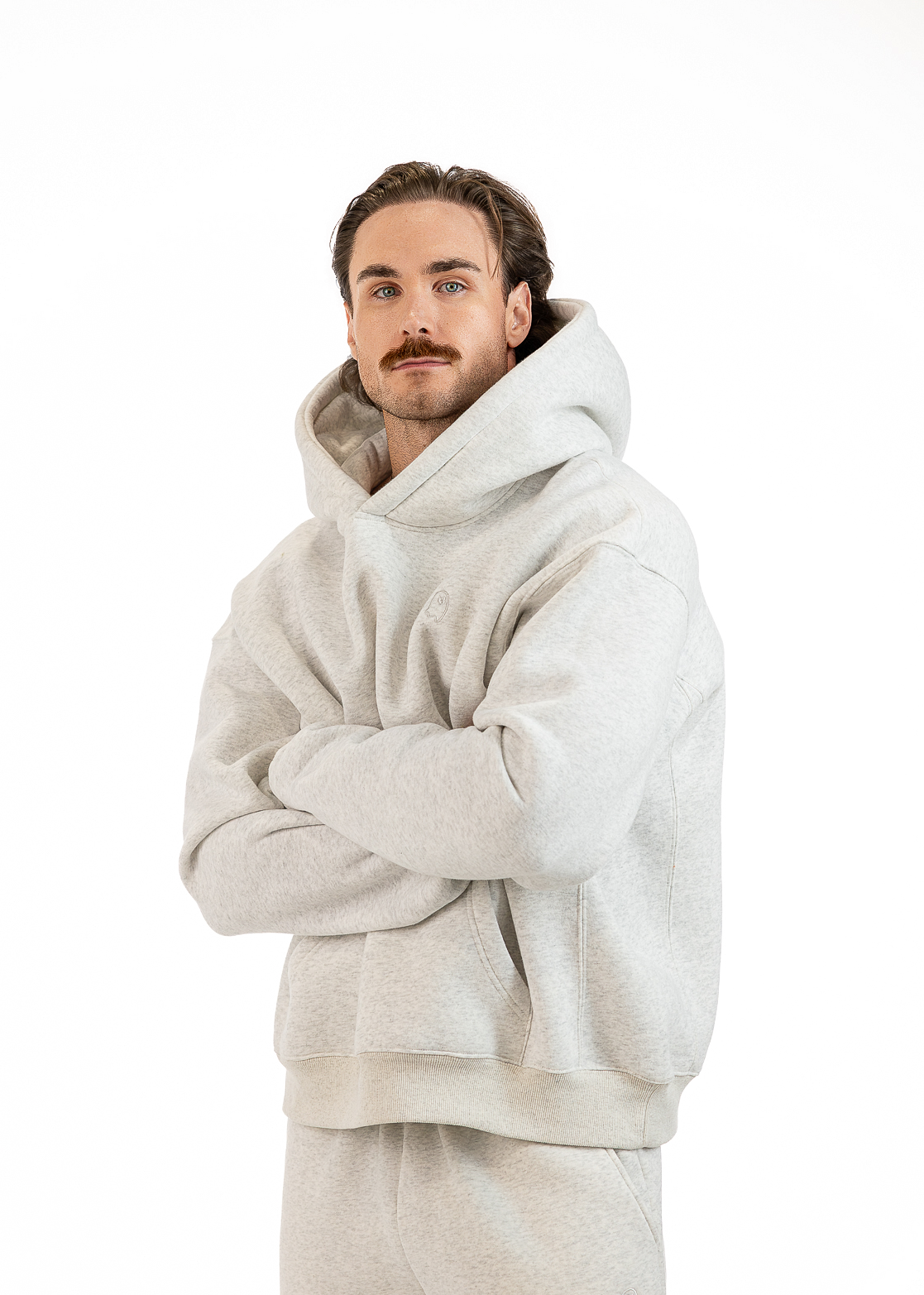 A man wearing a light gray oversized hoodie and matching sweatpants, standing with arms crossed against a plain white background.