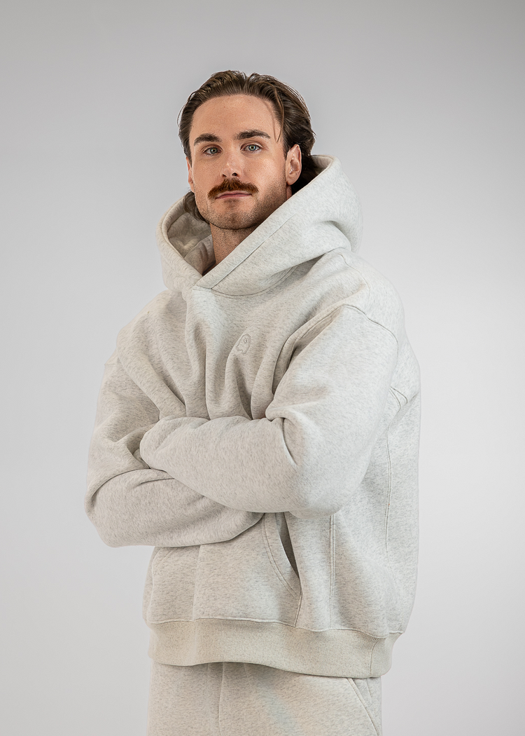A man wearing a light gray oversized hoodie and matching sweatpants, standing with his arms crossed against a neutral gray studio background.
