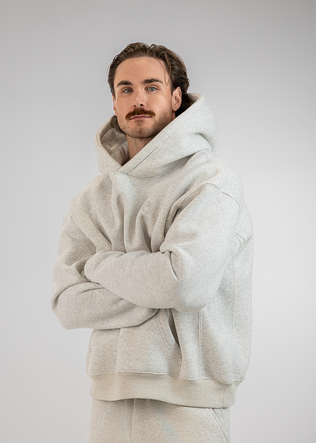 A man wearing a light gray oversized hoodie and matching sweatpants, standing with his arms crossed against a neutral gray studio background.
