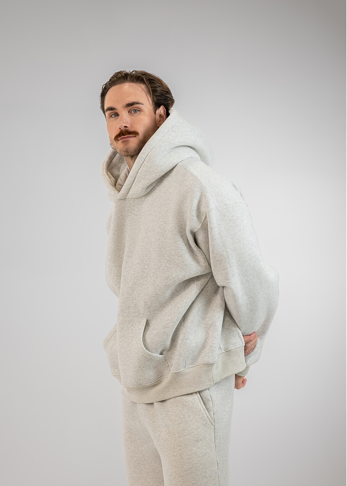 A man wearing a light gray oversized hoodie and matching sweatpants, photographed against a plain light gray studio background.
