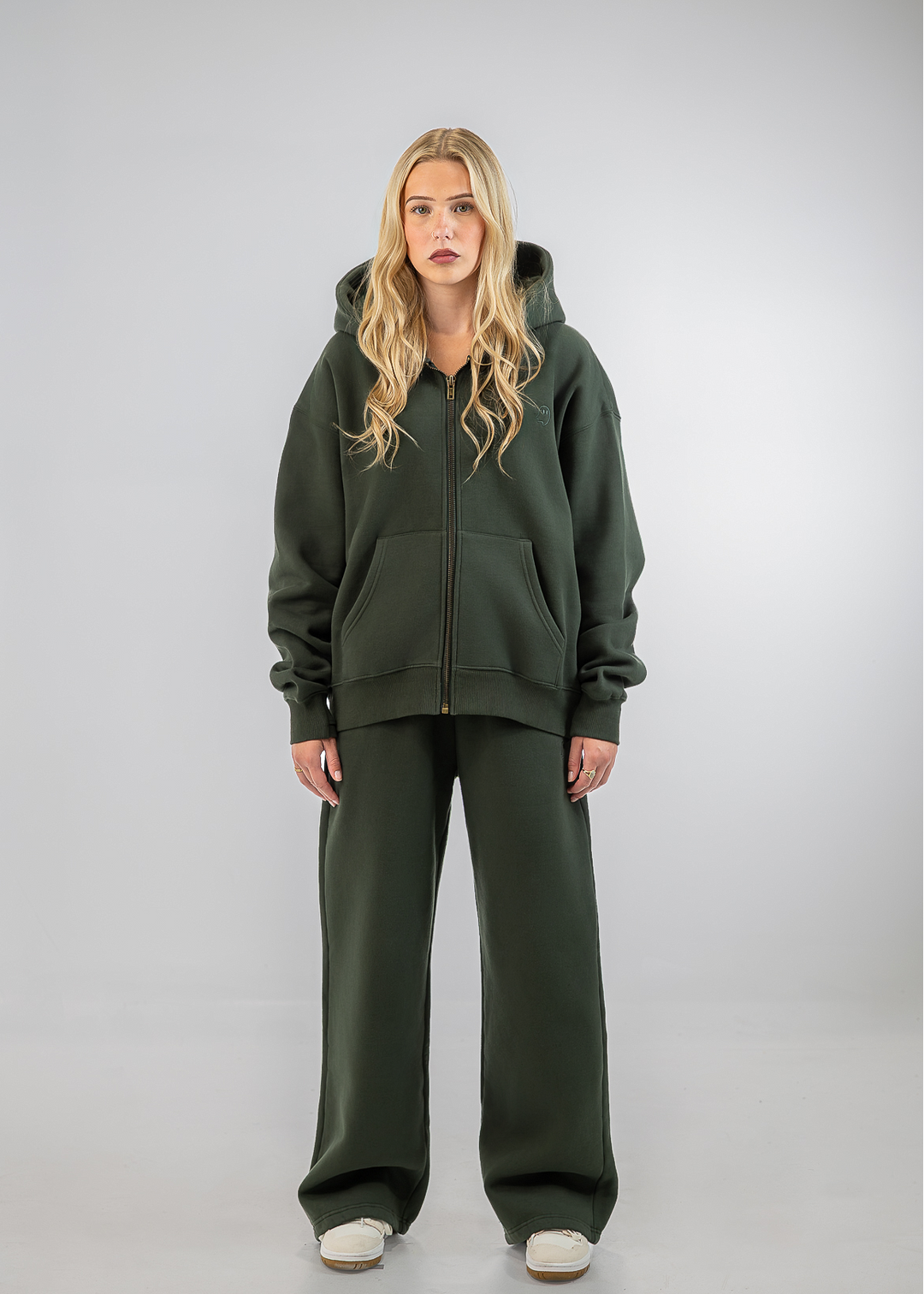 A woman modeling an oversized dark olive green zip-up hoodie and matching wide-leg sweatpants against a light gray studio background.